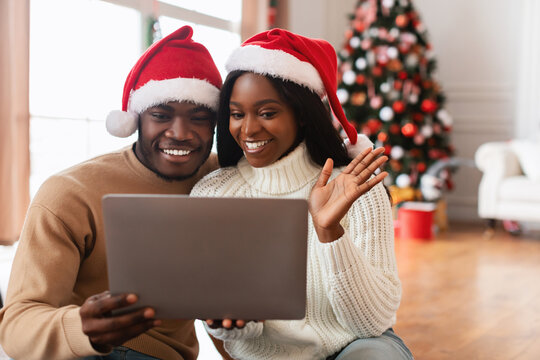 Cheerful black couple wearing red Santa hats enjoys a video call using a tablet. They smile and wave while sitting on the floor in a cozy living room decorated for Christmas.