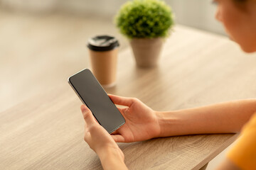Unrecognizable asian woman sits at a wooden desk holding a smartphone with a blank screen. She appears to be shopping online, with a coffee cup nearby and a small plant on the table.