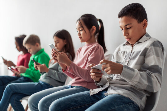 Group of diverse preteen children sit in a line against a gray background. Each child is focused on their smartphone, playing games and browsing social media feeds.