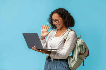A Black female student with curly hair and glasses waves at her laptop during an online class. She is wearing a casual outfit and has a backpack, engaged in a video chat with her college teacher.