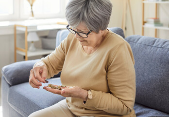 Portrait of poor old senior woman sitting on couch in the living room at home and counting coins in hands. Elderly person holding money in palms. Pension poverty and savings concept.