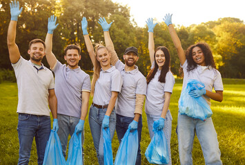 Happy volunteer group, friends standing happily in sunny green park, diverse team smiling, waving, holding bags and wearing gloves, feeling accomplished after volunteering and cleaning environment