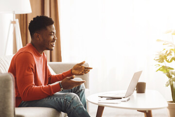 A young man wearing a red sweater is sitting on a couch, smiling and chatting during a video call on his laptop. Sunlight fills the room, creating a warm atmosphere.