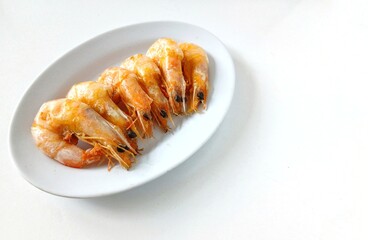 A minimalist food photograph featuring six boiled shrimp neatly arranged in a row on a simple white oval plate. The image is shot from above against a clean white background, highlighting the natural.