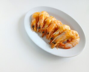 A minimalist food photograph featuring six boiled shrimp neatly arranged in a row on a simple white oval plate. The image is shot from above against a clean white background, highlighting the natural.