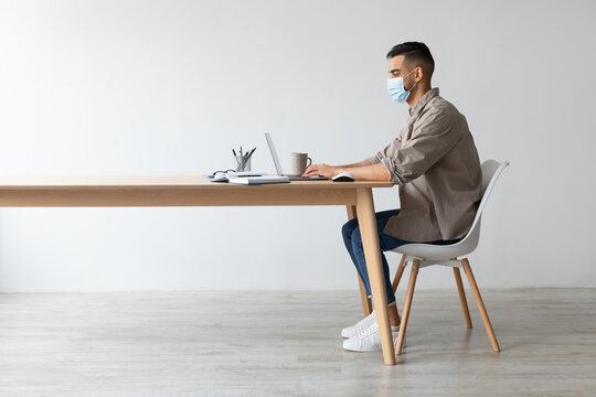 A young business man is focused on his laptop at a home office. He wears a disposable facemask while sitting at a table. This scene reflects the reality of working from home during quarantine.