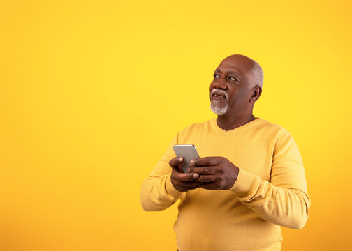 Cheerful senior African American man is using a mobile phone, exploring a new application. He looks engaged while standing against a bright orange background, enjoying modern technology.