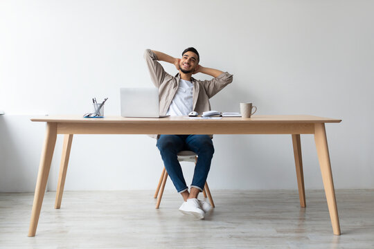 A smiling Arab man relaxes in his chair at a desk, taking a break from work while using his laptop. He leans back with his hands behind his head, enjoying a moment of downtime.