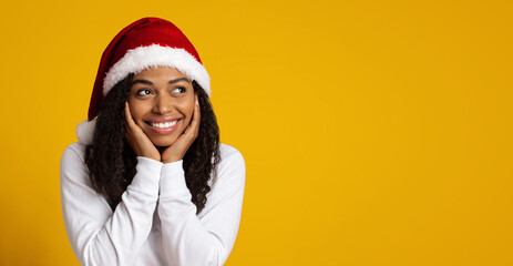 A joyful young woman wearing a Santa hat smiles widely while resting her hands on her cheeks. The bright yellow background adds a festive atmosphere perfect for the holidays.