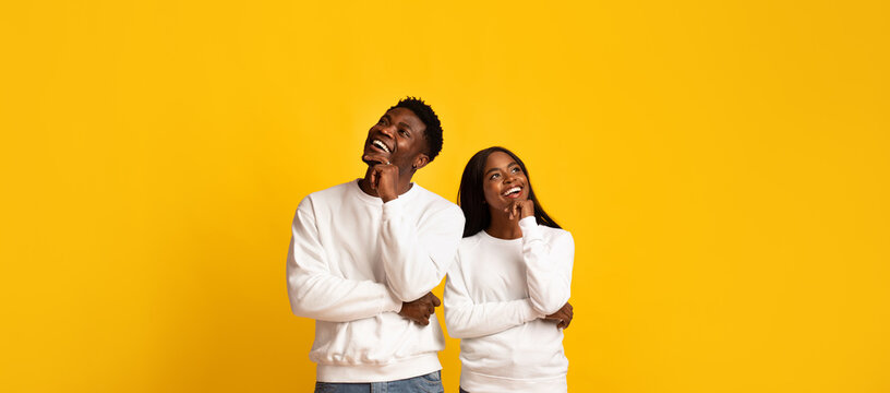 A happy couple stands side by side, both wearing white sweaters and smiling thoughtfully. They gaze upward against a vibrant yellow background, exuding joy and connection.