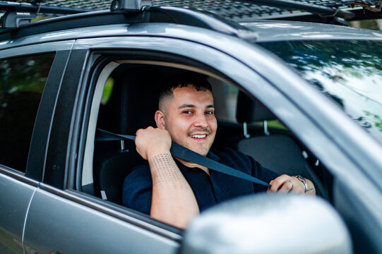 An adult man in his thirties is smiling while fastening his seatbelt in a parked car.