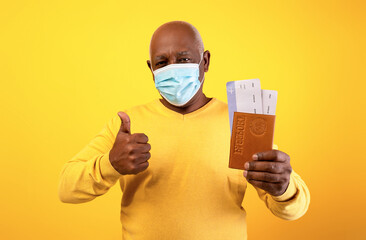 Senior African American man shows excitement for travel while holding his passport and tickets. He wears a face mask for safety against covid-19, showcasing positivity in challenging times.