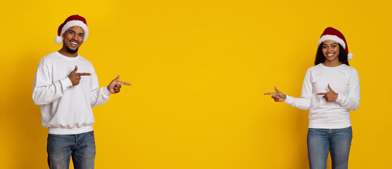 Two happy individuals wear Christmas hats and point joyfully to their sides in front of a vibrant yellow wall. Their cheerful expressions enhance the festive atmosphere.