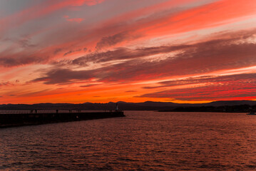 Naklejka premium A vibrant sunset over the Pacific coast, the sky is saturated with vibrant colors, the horizon is marked by distant hills. The composition is serene and dramatic due to the contrast and natural beauty