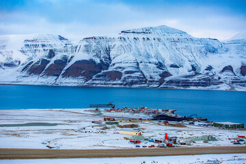 Longyerbeyen, Svalbard, Snowy mountain range with a small town in the valley below. The town is surrounded by snow and the water is calm. The scene is peaceful and serene