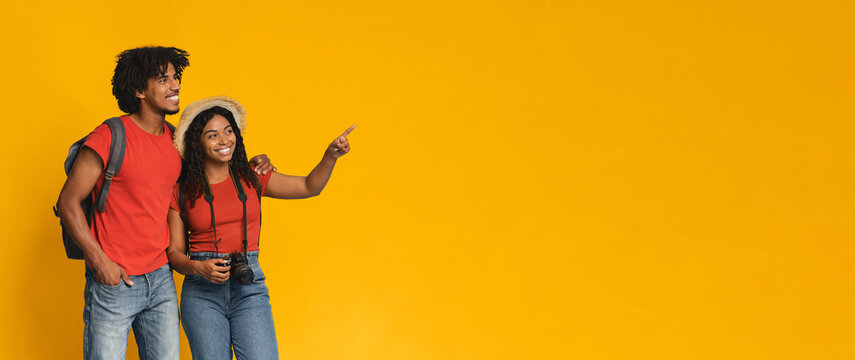 A happy couple stands together in front of a bright yellow wall. They smile, with one pointing excitedly, and the other holds a camera, ready to capture the moment.