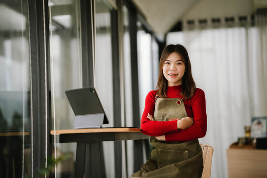 A smiling barista in a red sweater and apron holds a tablet while standing inside a cozy coffee shop. - Powered by Adobe