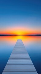 Obraz premium Wooden pier stretches across calm water towards vibrant orange sunset reflecting on the glassy surface under a clear blue sky