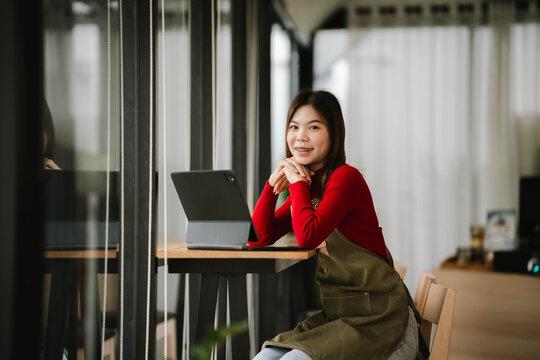 A smiling barista in a red sweater and apron holds a tablet while standing inside a cozy coffee shop. - Powered by Adobe