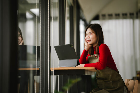 A smiling barista in a red sweater and apron holds a tablet while standing inside a cozy coffee shop. - Powered by Adobe