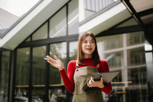 A woman wearing an apron holds a tablet while working outdoors in front of a modern building.