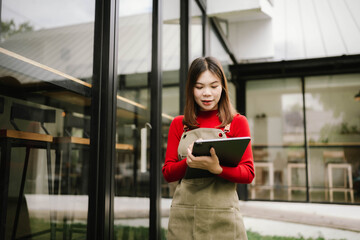 A woman wearing an apron holds a tablet while working outdoors in front of a modern building.