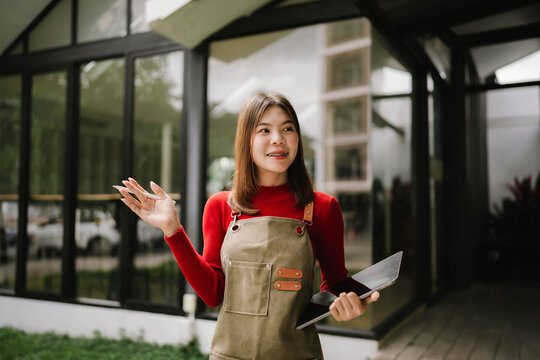 A woman wearing an apron holds a tablet while working outdoors in front of a modern building.