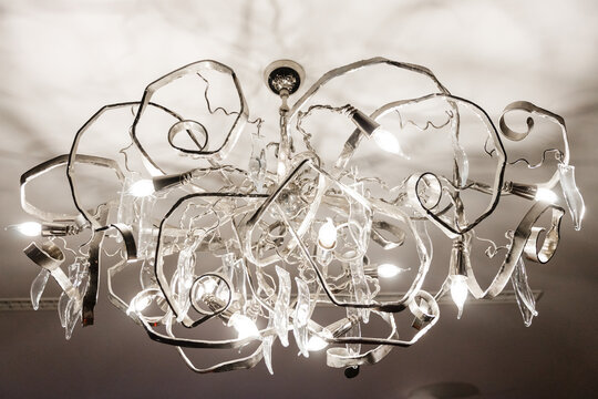 Striking modern chandelier with multiple glowing bulbs and abstract, organic-shaped glass and metal elements, providing bright decorative light against a dark ceiling