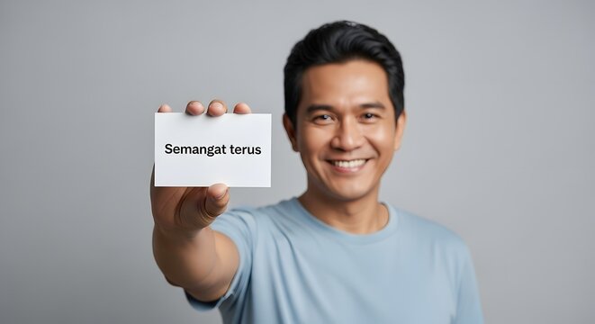 A smiling man holding a sign with the message 'Semangat terus' in front of a plain gray background, conveying motivation and positivity