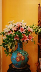 A stunning bouquet of pink and white lilies bursts from an ornate turquoise vase, decorated with peach motifs, set against a warm yellow wall inside the Penang Peranakan Mansion.