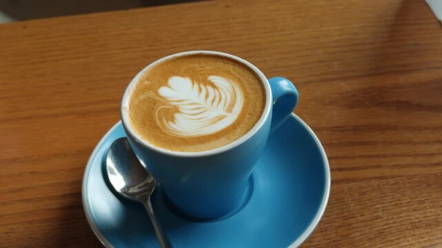 A close-up shot of a skilled barista creating beautiful latte art, specifically a heart design, with steamed milk into a blue ceramic coffee cup. The scene is set on a rustic wooden table, emphasizing