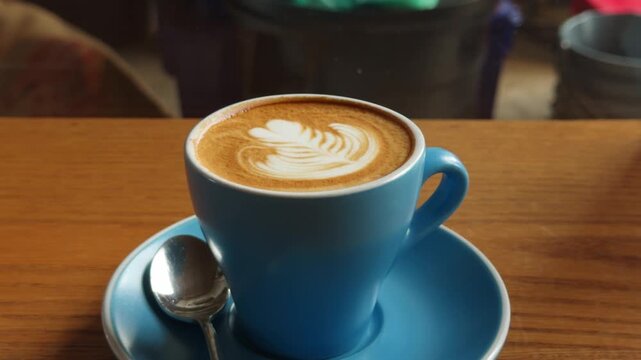 A close-up shot of a skilled barista creating beautiful latte art, specifically a heart design, with steamed milk into a blue ceramic coffee cup. The scene is set on a rustic wooden table, emphasizing