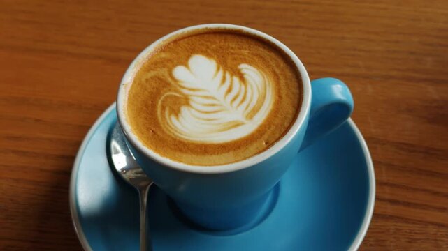 A close-up shot of a skilled barista creating beautiful latte art, specifically a heart design, with steamed milk into a blue ceramic coffee cup. The scene is set on a rustic wooden table, emphasizing
