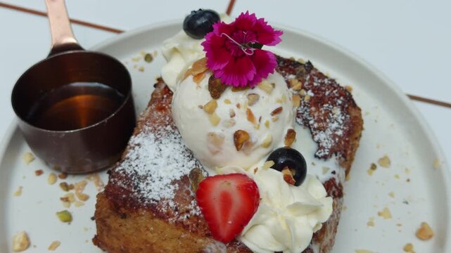 A close-up and highly appetizing shot of a gourmet French toast dessert, artfully plated on a white ceramic dish. The thick-cut toast is generously topped with fluffy whipped cream, a colorful assortm