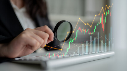 A close-up view of hands using a magnifying glass over financial graphs on a keyboard, illustrating...