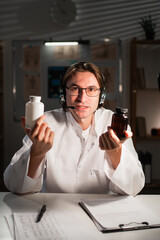 Doctor recommending drug, pills and medicine. Young therapist in headphones and eyeglasses showing jar of pills looking at camera during night shift in hospital