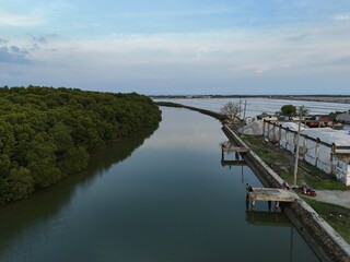 Obraz premium Aerial view of a serene river separating a lush mangrove forest from an industrial coastal area under an overcast sky