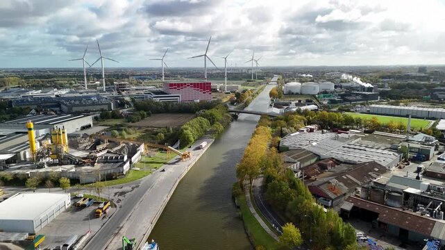 Aerial view of an industrial area with wind turbines and a canal running through it. The scene shows renewable energy integration in a modern European industrial landscape.