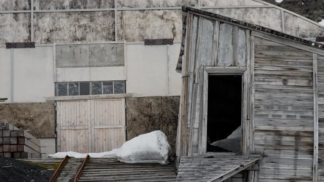 Weathered wooden outbuilding with an open doorway and snowbank set against a stark industrial wall, featuring peeling planks, muted winter textures, and signs of decay and neglect