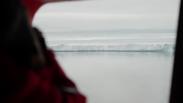 Passenger's window view of a solitary tabular iceberg drifting across a calm sea under an overcast sky, with a blurred red jacket in the foreground framing the tranquil seascape.