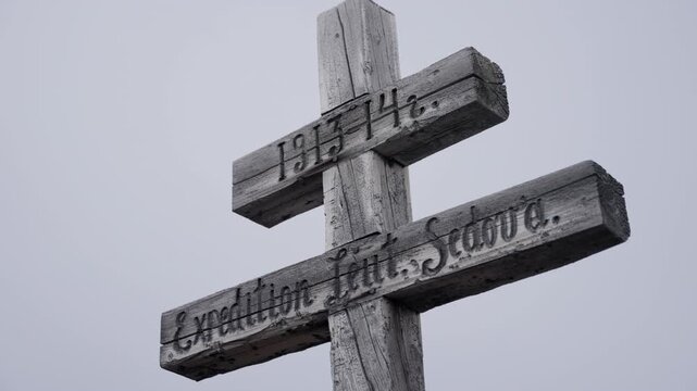 Weathered double-barred wooden memorial cross with carved inscription and 1913 14 dates, standing on a barren hillside beneath an overcast sky, signifying remembrance.