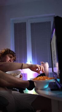 Vertical screen single man eats snacks with his legs up while watching evening content in a neon lit room creating a relaxed carefree atmosphere that reflects simple bachelor habits and laid back nigh