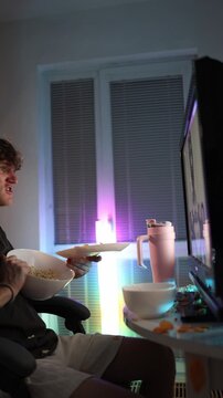 Vertical screen young man throws chips and popcorn at monitor showing frustration anger and emotional reaction to disappointing video content during late night entertainment under neon light