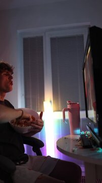 Vertical screen angry young man throws popcorn and chips at monitor showing irritation and emotional reaction to bad content during night entertainment under colorful neon light in messy room