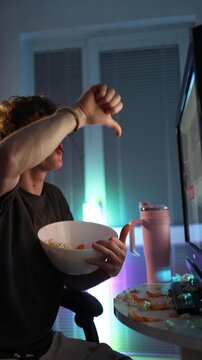 Vertical screen young man furiously throws popcorn and chips at monitor expressing frustration anger and emotional reaction to bad content during night entertainment in messy colorful room