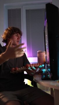 Vertical screen young man furiously throws eggs at monitor reacting to terrible movie scene expressing frustration and disappointment through messy impulsive act under colorful neon light at home