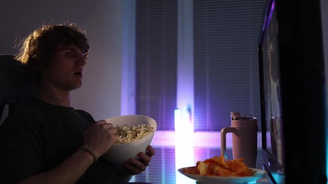 Young man watches the screen at night with a bowl of popcorn reacting with surprise and disbelief neon light fills the room creating a simple relatable moment of unexpected emotion during quiet home v