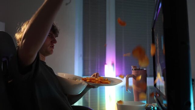 Young man throws chips at the monitor in frustration reacting to disgusting online content neon light fills the room creating a chaotic messy moment driven by irritation and impulsive emotion at night