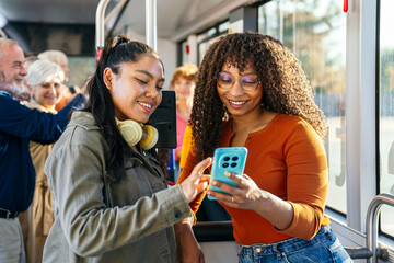 Two smiling multi ethnic women friends are sharing a smartphone while traveling on a city public bus, representing urban commute and connection