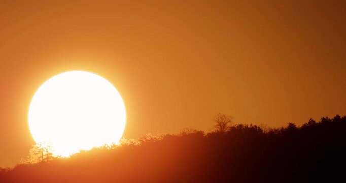 Sunrise time lapse, massive bright orange sun rising rapidly over a dark mountain forest silhouette catching the vivid morning golden hour 4K